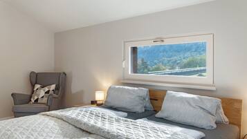 A bedroom with a bed, a chair, a bedside table with a lamp, and a window with a view of trees and a bridge.