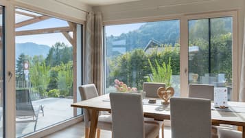 Área de refeições com mesa, cadeiras, janelas amplas e vista para vegetação e colinas.