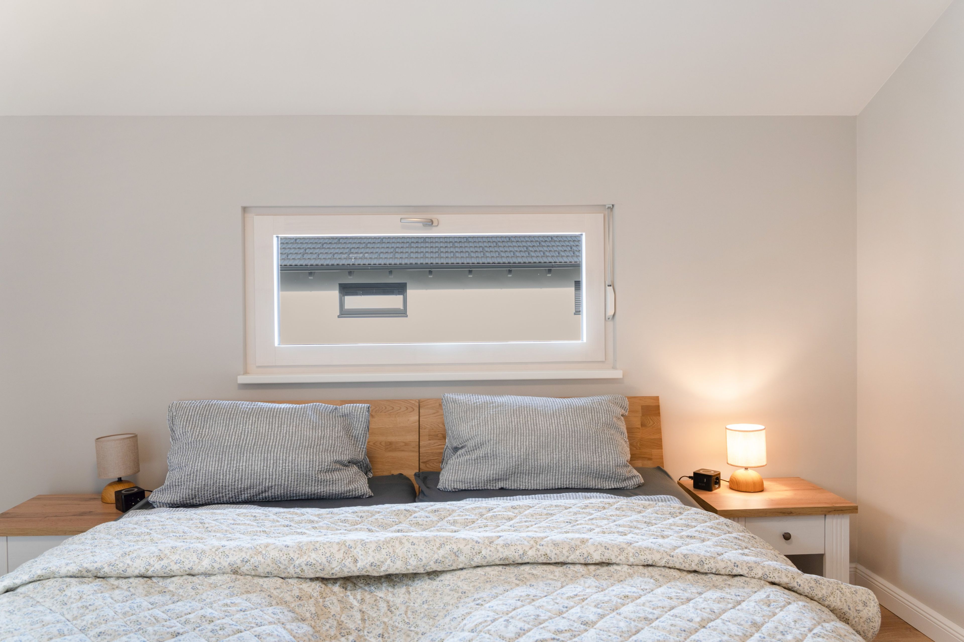 A bedroom with a bed, two bedside tables, a nightstand, a lamp, a window, and a wall-mounted shelf.