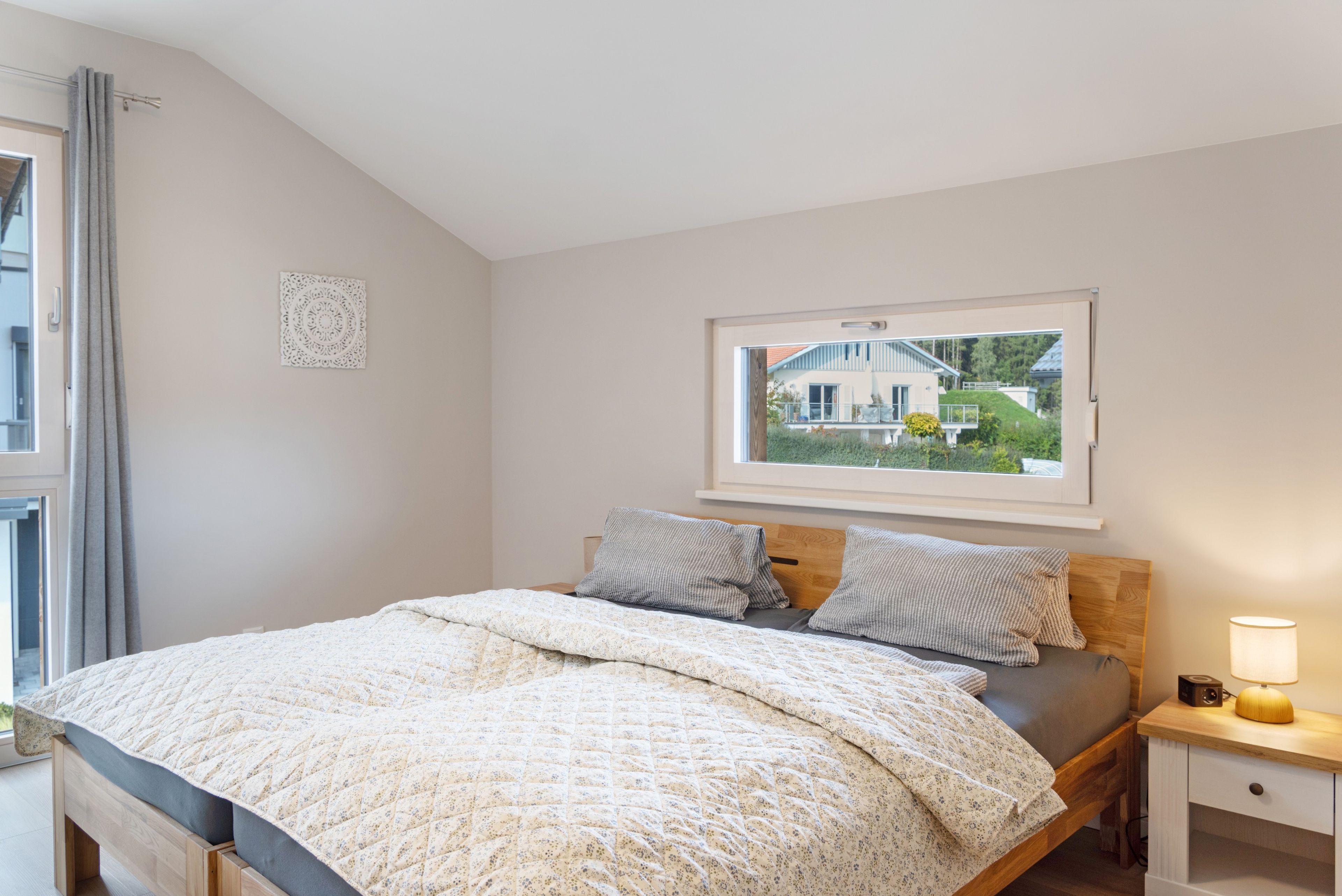 Quarto com cama, mesa de cabeceira, luminária e janela com vista para uma casa.