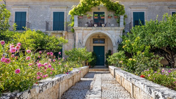 A building with a central entrance, a balcony with plants, and a stone pathway leading to the entrance. - Villa Aguglie - Suite Dalia (Modica)