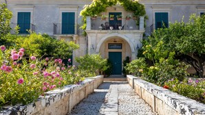 A building with a stone pathway leading to a green door, surrounded by flowering plants and greenery. - Villa Aguglie - Suite Dalia (Modica)