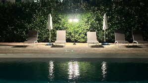 A poolside area with lounge chairs and umbrellas, a building with a tiled roof in the background, and a hedge in the foreground. - Villa Aguglie - Suite Dalia (Modica)