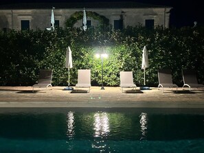 A poolside area with lounge chairs and umbrellas, a building with a tiled roof in the background, and a hedge in the foreground. - Villa Aguglie - Suite Rosa (Modica)