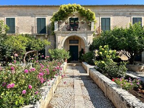 A courtyard with a stone pathway leading to a building with a green door and a balcony with plants. - Villa Aguglie - Suite Rosa (Modica)