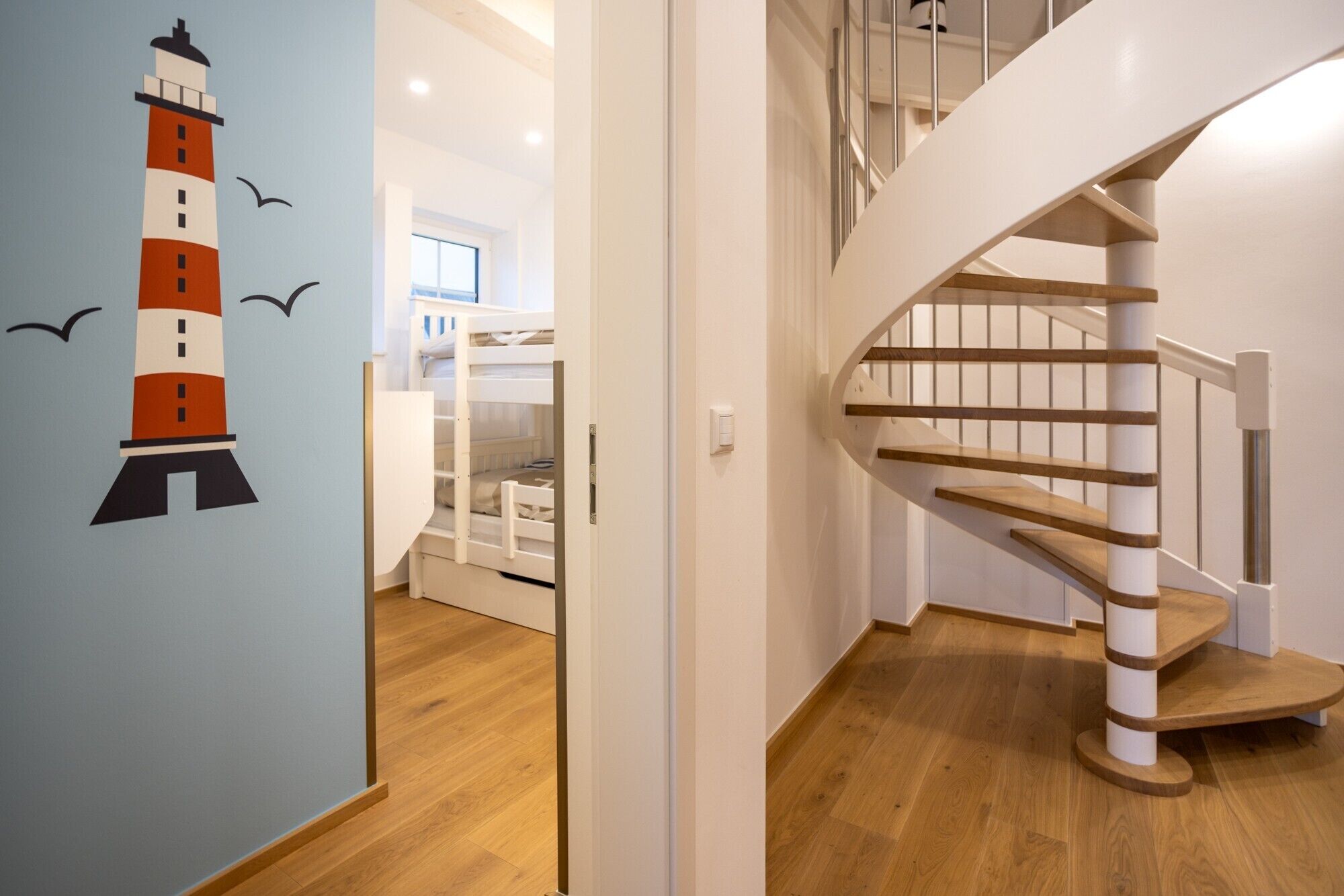 Un escalier en colimaçon avec des marches en bois et une rampe blanche, menant à une pièce où se trouve un lit superposé et une fenêtre.