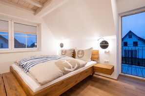 A bedroom with a wooden bed, a nightstand, a wall-mounted lamp, and a window with a view of a blue house.