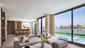 A modern living room with a large window overlooking a pool and a view of buildings. There is a sofa, a coffee table, and a bar area with stools. - Villa Portofino by Luxury Explorers Collection (Dubai)