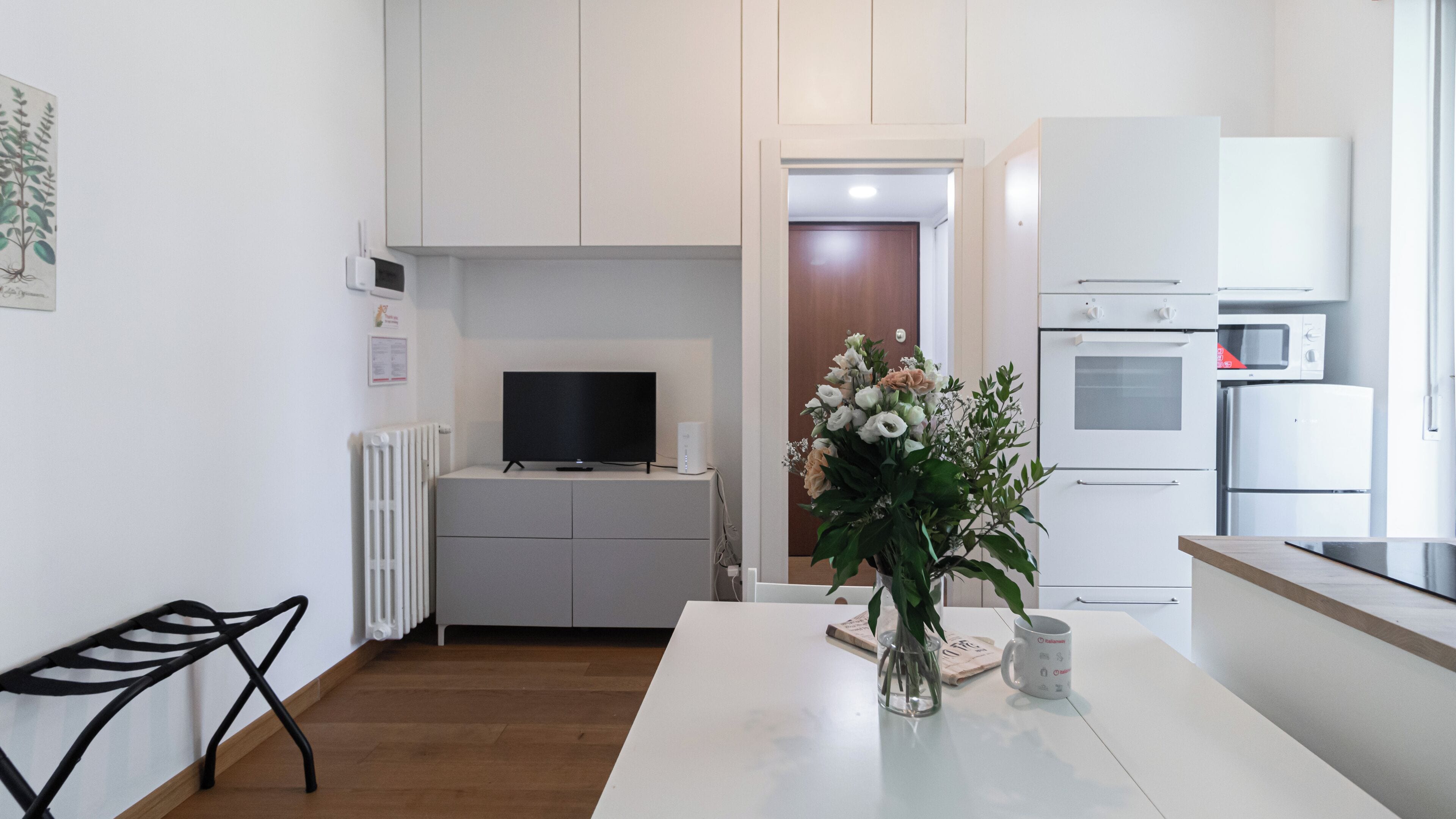 A modern kitchen with white cabinets, a built-in oven, microwave, and refrigerator. There's a dining table with a vase of flowers and a coffee mug.