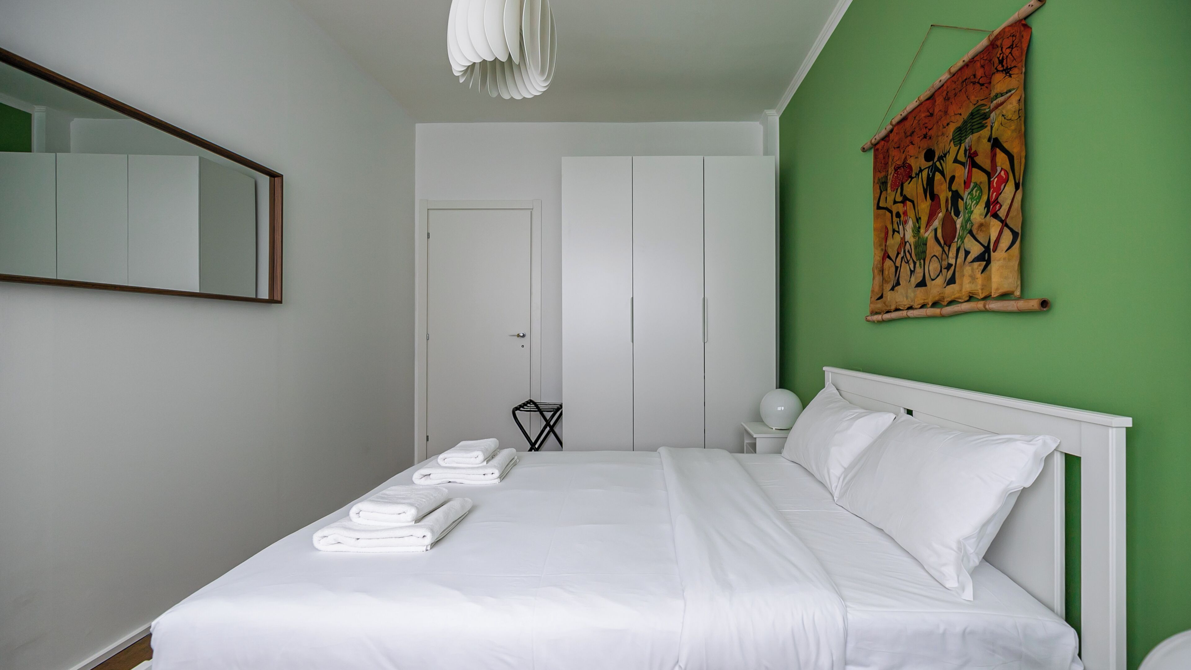 A neatly made bed with white linens and towels, a white nightstand with a lamp, a mirror on the wall, and a green wall with a tapestry.