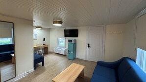 A compact living space with a wooden floor, a blue sofa, a wooden table, a kitchenette with a fridge and microwave, a TV mounted above the fridge, and a door with a handle.