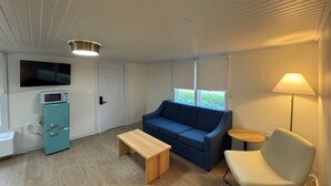 A compact living space with a blue sofa, a wooden table, a white armchair, a small lamp, a microwave, and a TV on the wall.