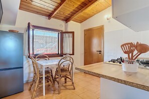 A kitchen with a wooden ceiling, a dining table with chairs, a refrigerator, a window with curtains, and a door.