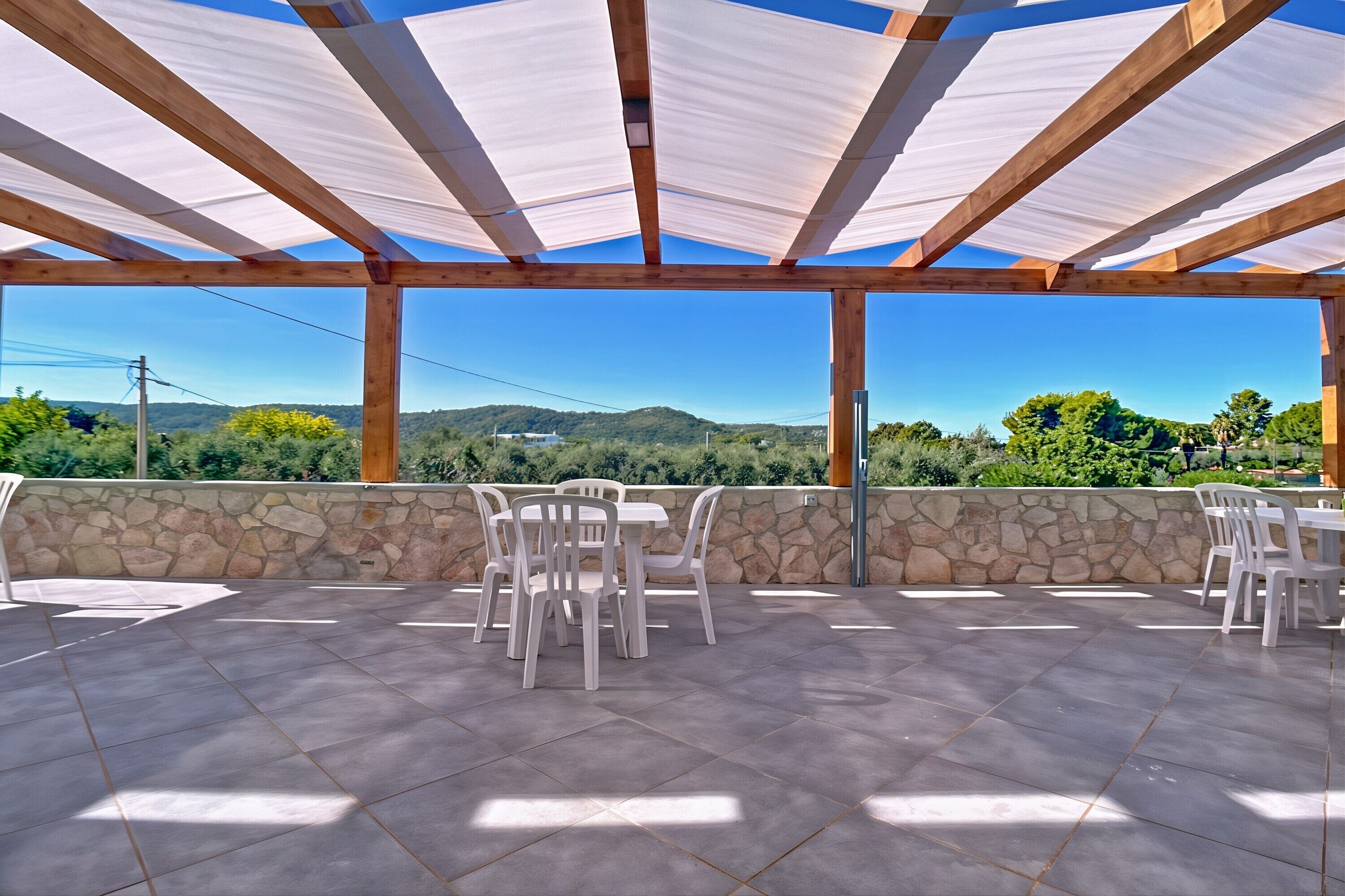 An outdoor patio with stone walls, a wooden pergola, and white plastic chairs.