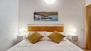 Ein ordentlich bezogenes Bett mit zwei Kissen, ein Gemälde einer Küstenlandschaft und zwei Nachttischlampen.