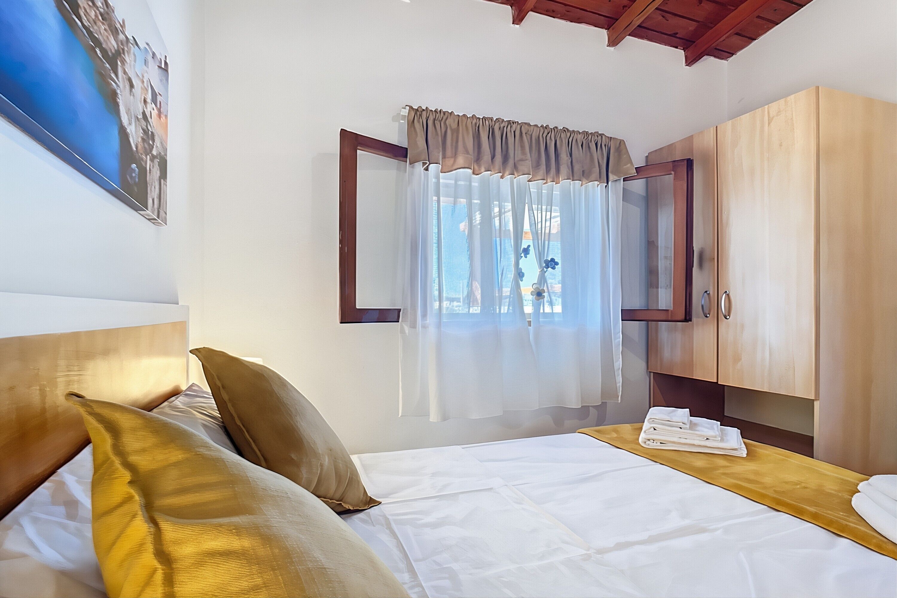 A hotel room with a bed, a wooden wardrobe, a window with sheer curtains, and a painting on the wall.