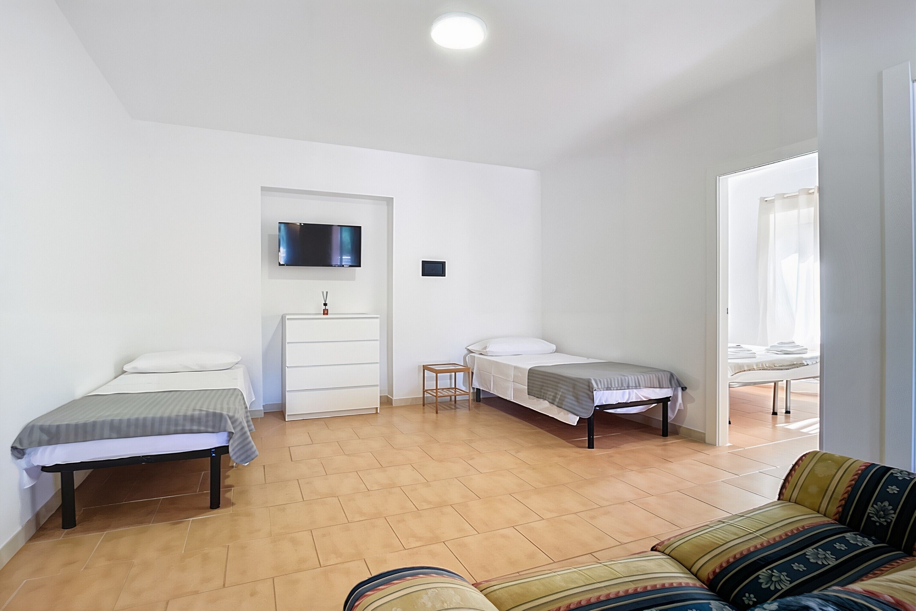 Un dormitorio con dos camas individuales, una mesita de noche pequeña, un televisor montado en la pared y una ventana con cortinas.