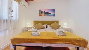 Un lit recouvert d’une couvre-lit jaune, avec deux oreillers, et un tableau représentant un paysage côtier accroché au-dessus.