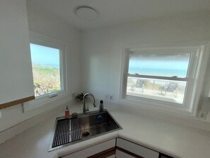 A kitchen with a stainless steel sink, two windows with views of the beach, and a ceiling vent.
