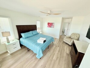 A bedroom with a bed, bedside table, lamp, chair, and a ceiling fan. - Salty Haven - Endless Views - Beachfront & Pool - 7BR (Indian Beach)