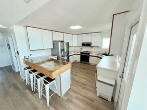 A modern kitchen with white cabinetry, a central island with stools, stainless steel appliances, and a herringbone floor pattern. - Salty Haven - Endless Views - Beachfront & Pool - 7BR (Indian Beach)