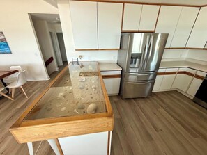 A modern kitchen with a glass-top island, stainless steel appliances, and wooden flooring. - Salty Haven - Endless Views - Beachfront & Pool - 7BR (Indian Beach)