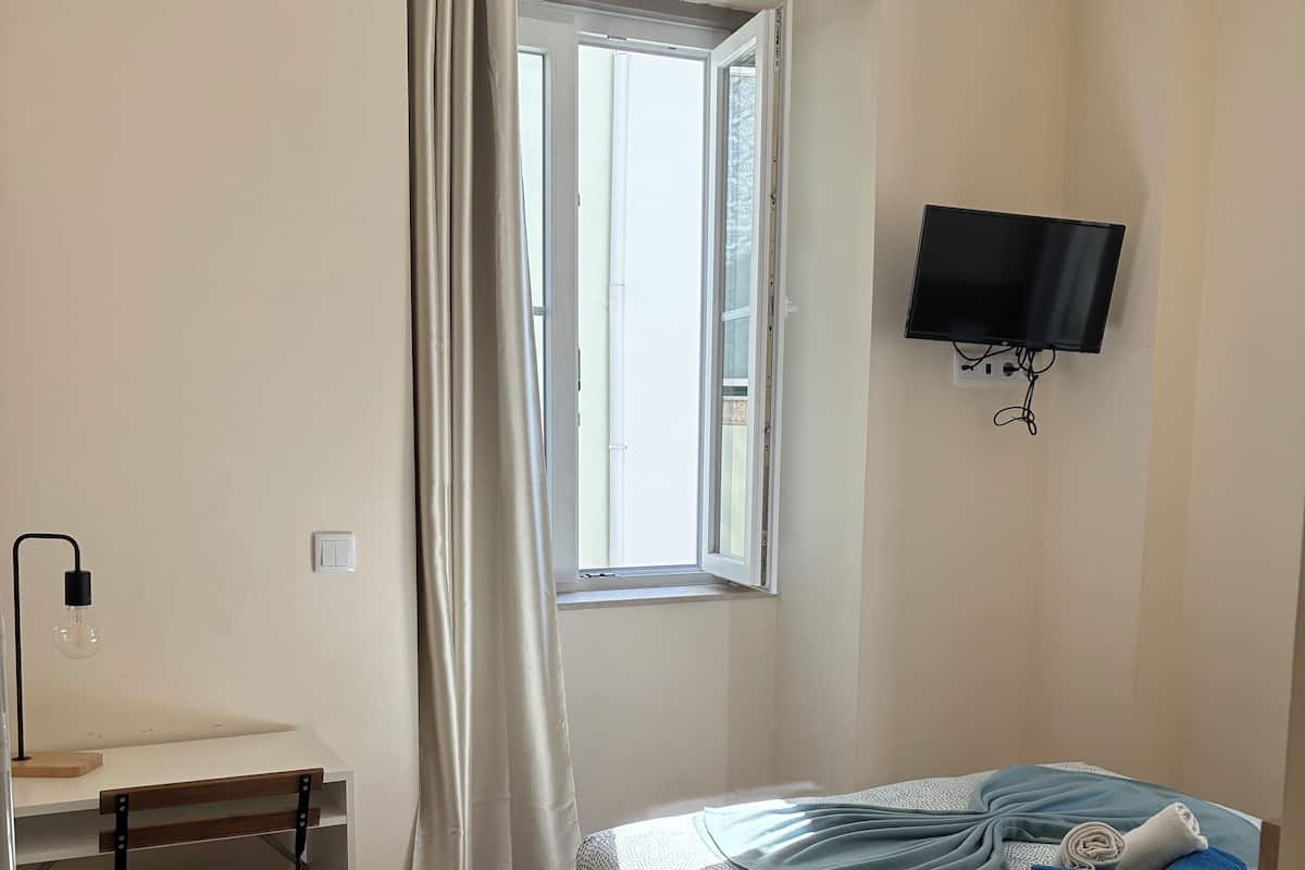 A bedroom with a bed, a desk, a chair, a TV, and a window with curtains.
