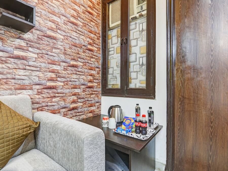 A room with a brick wall, a window with patterned glass, a sofa with a cushion, and a table with a kettle, cups, and snacks.