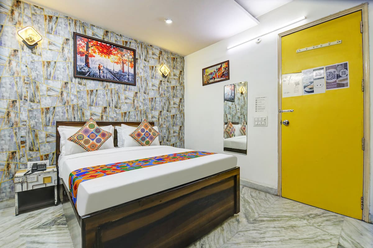 Una habitación de hotel con cama, mesita de noche, espejo, puerta amarilla y un papel tapiz estampado en la pared.