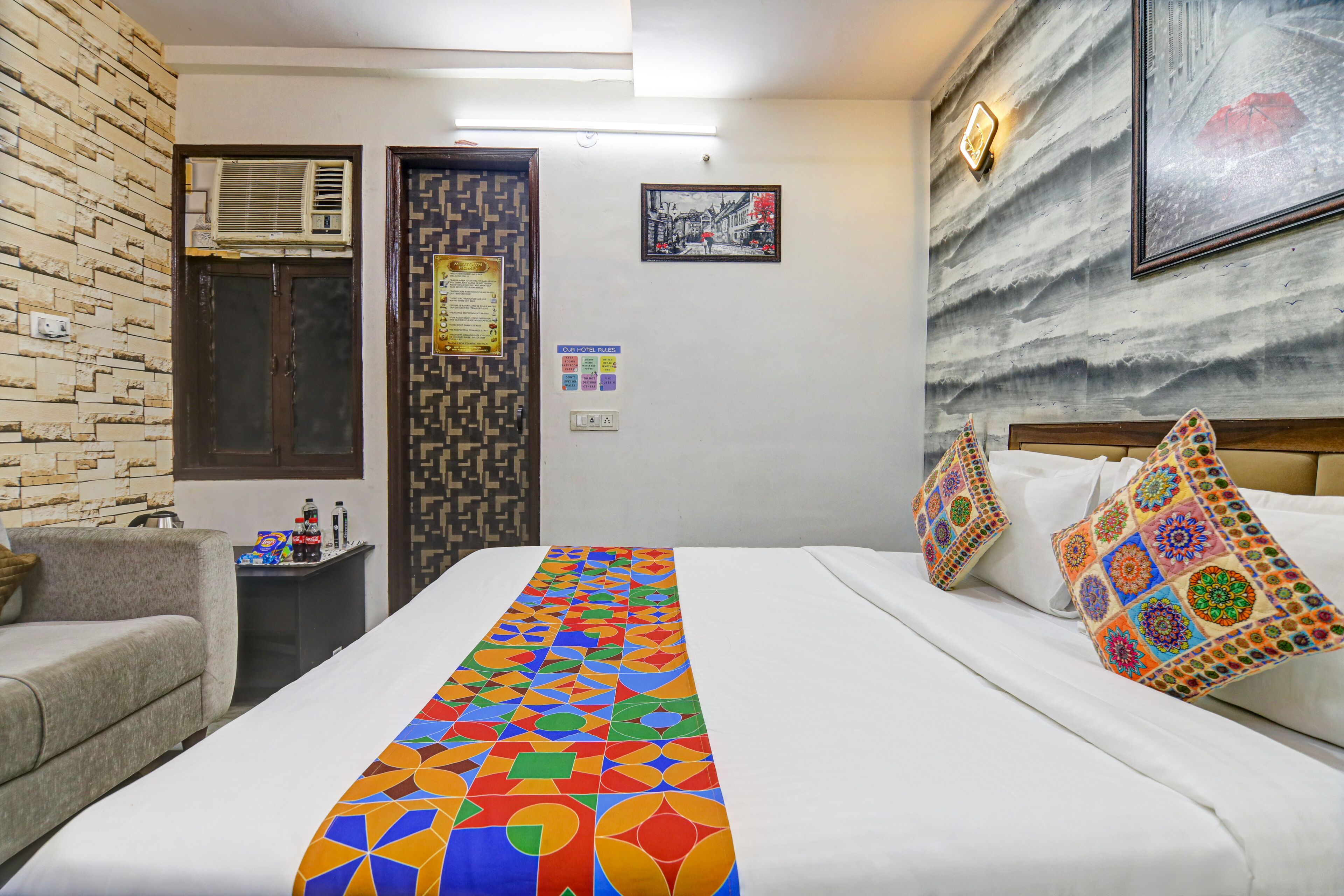 Une chambre d’hôtel comprenant un lit, un canapé, une petite table avec quelques objets, un climatiseur fixé au mur et une porte ornée d’un motif décoratif.