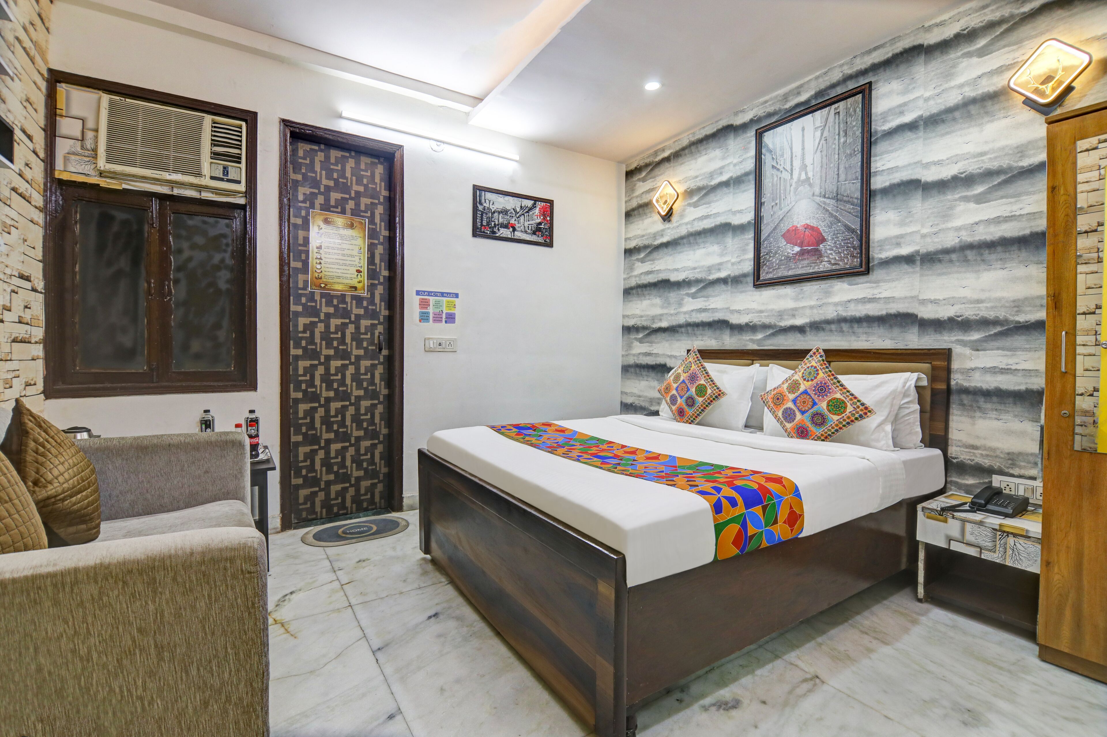 A hotel room with a bed, a sofa, a nightstand, a wall-mounted air conditioner, a patterned door, and a framed picture on the wall.