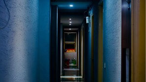 Un couloir étroit, avec un mur bleu d’un côté et une porte en bois de l’autre. Un luminaire est fixé au plafond et une poubelle se trouve au centre.