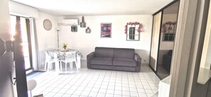 A living room with a gray sofa, a small round table with a vase of yellow flowers, a white tiled floor, and a large window with pink floral decorations.