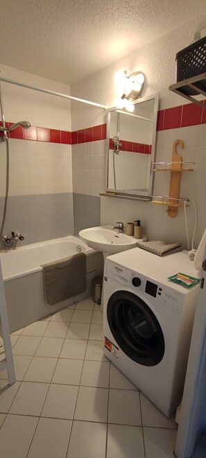 A compact bathroom with a white sink, a mirror, a shower, and a front-loading washing machine.