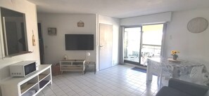 A living room with a flat-screen TV, a microwave, a white cabinet, a small table with a lamp, and a balcony with a table and chairs.