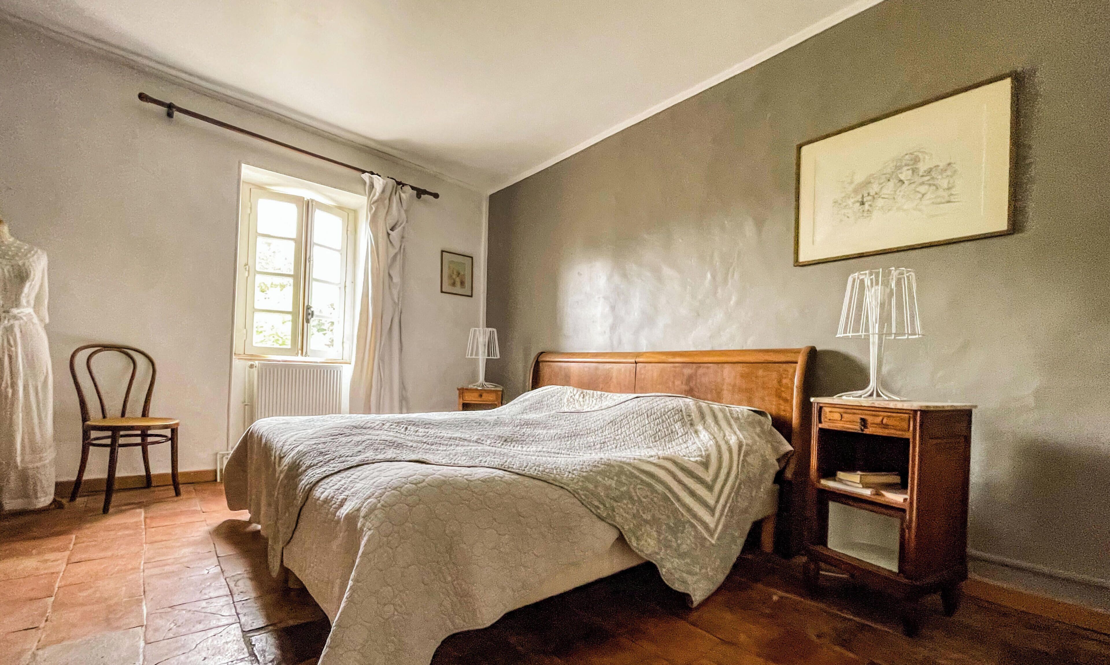 A bedroom with a bed, a chair, a nightstand, a lamp, a window, and a painting on the wall.