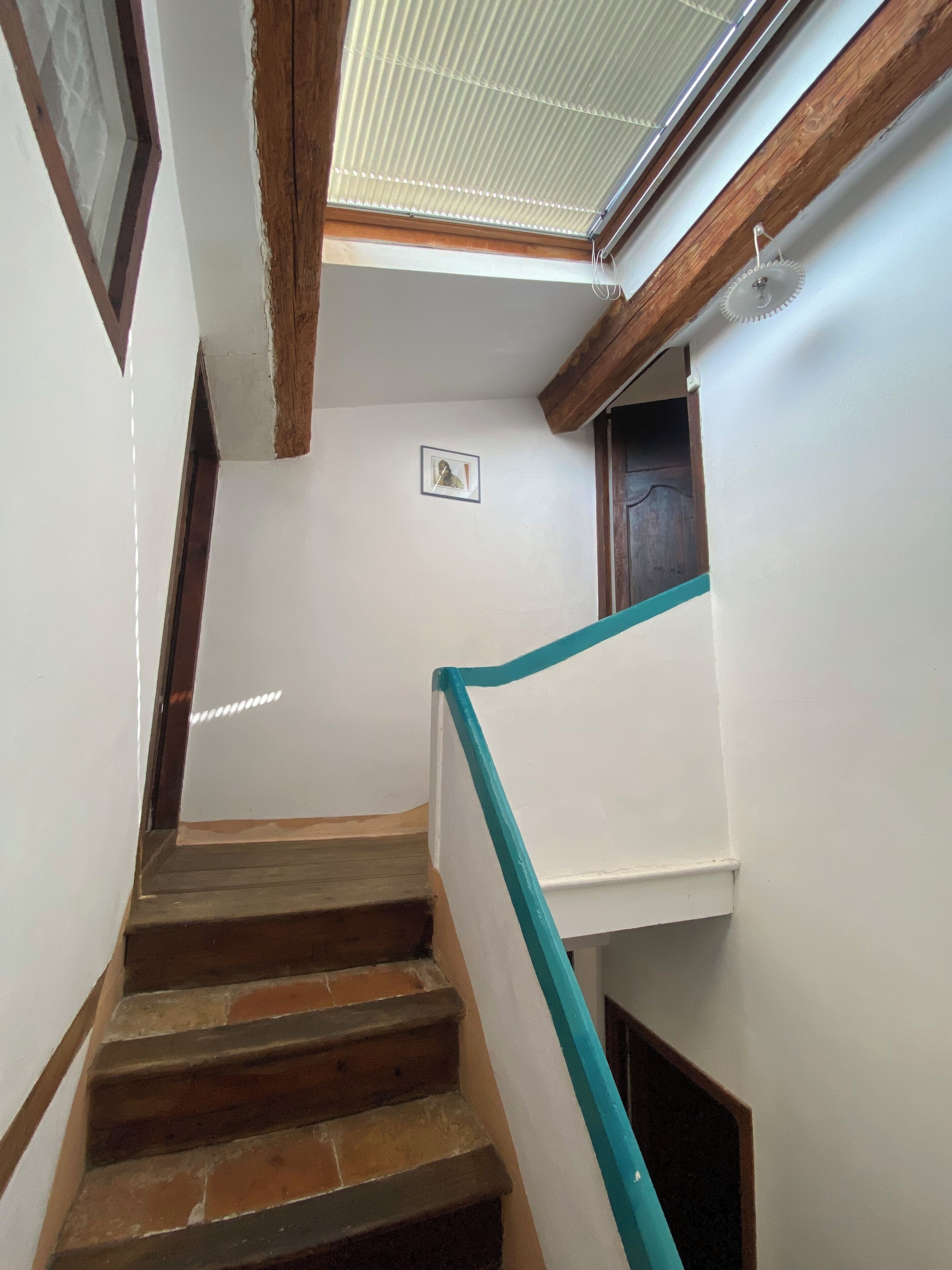 Un escalier avec des marches en bois et une rampe bleue, menant à une pièce dotée d’une fenêtre et d’un tableau encadré au mur.