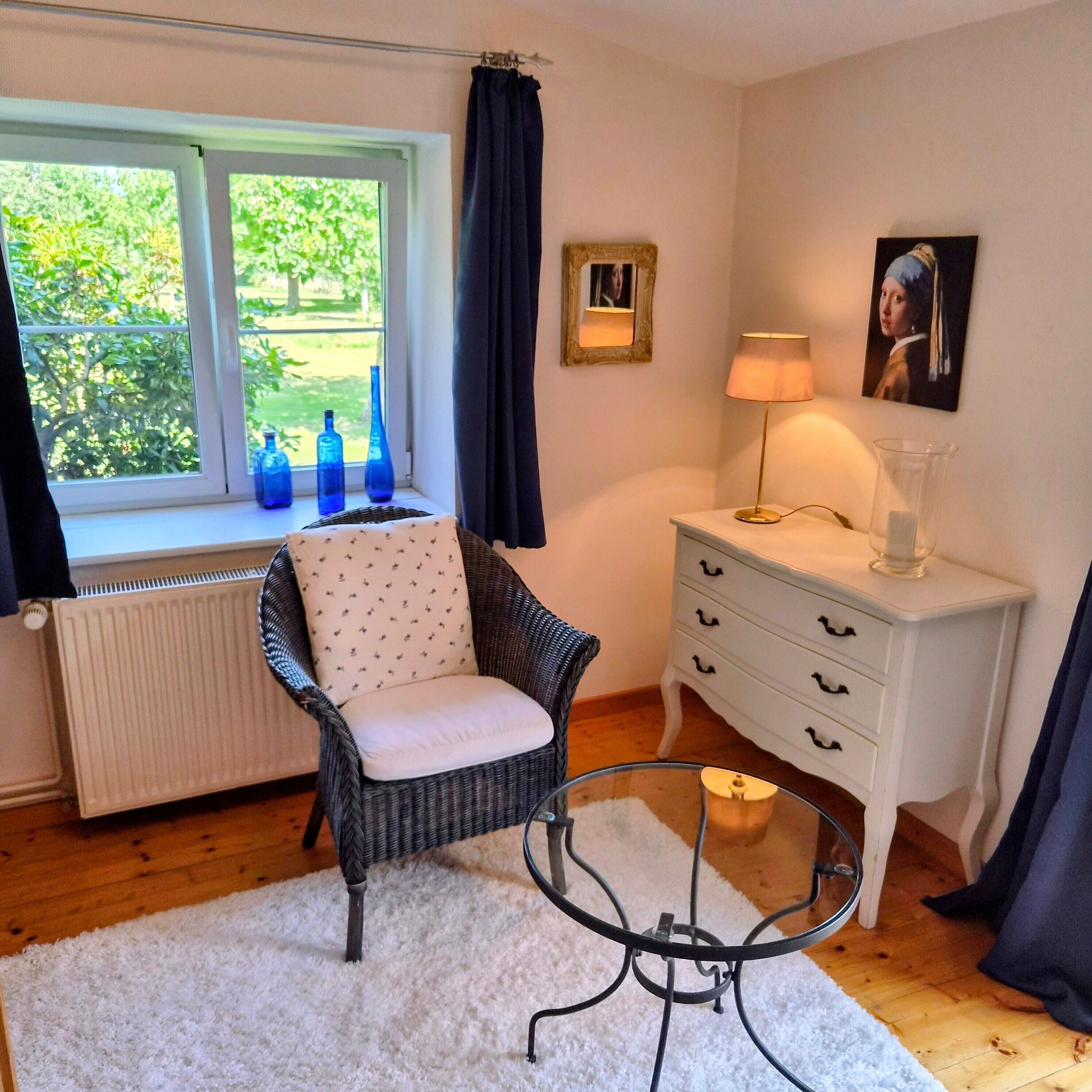 A room with a wicker chair, a white dresser, a glass table, and a window with blue vases on the sill.
