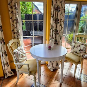 A room with a round table and two chairs, a window with curtains, and a view of a garden.