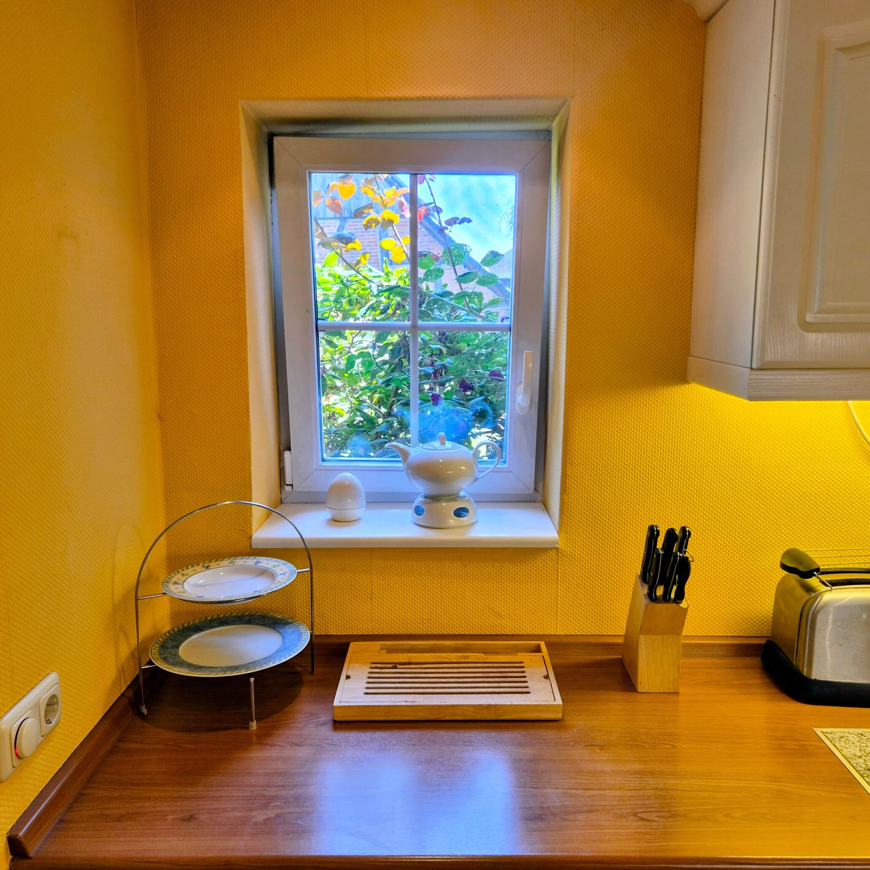 A small kitchen with a window, a tea set, a toaster, and a knife block.