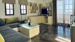 A modern living room with a sofa, a glass coffee table, a flat-screen TV, and built-in shelving. - Desire Apartments (Kato Gouves)