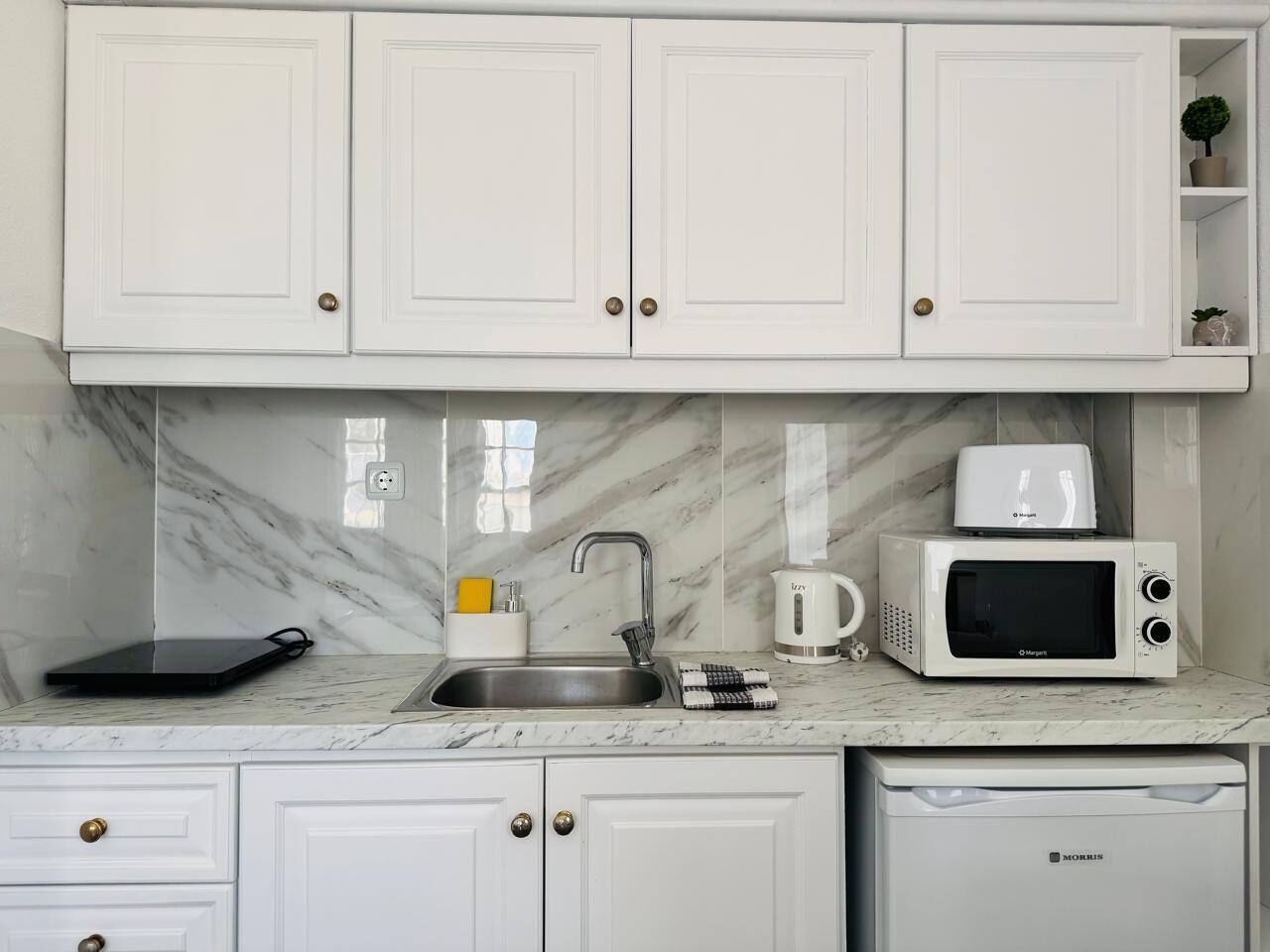 A kitchen with white cabinets, a marble countertop, a sink, a microwave, and a kettle.