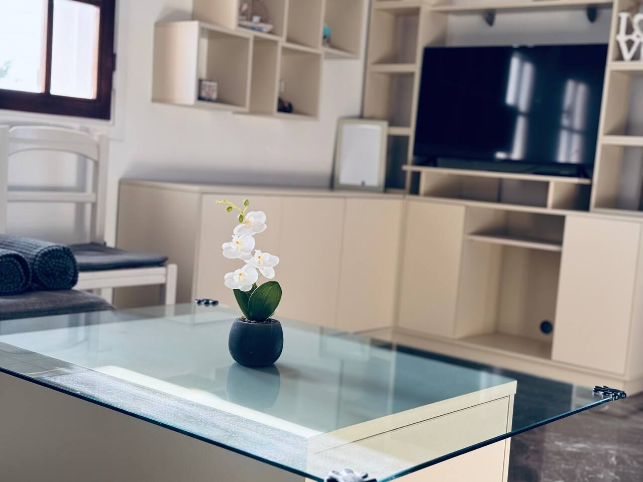 Una sala de estar moderna con una mesa de centro de vidrio, una orquídea en maceta pequeña y un televisor al fondo.