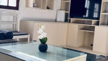Una sala de estar moderna con una mesa de centro de vidrio, una orquídea en maceta pequeña y un televisor al fondo.