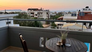 A balcony with a round wooden table and a chair, overlooking a residential area with buildings and a sunset view. - Desire Apartments (Kato Gouves)