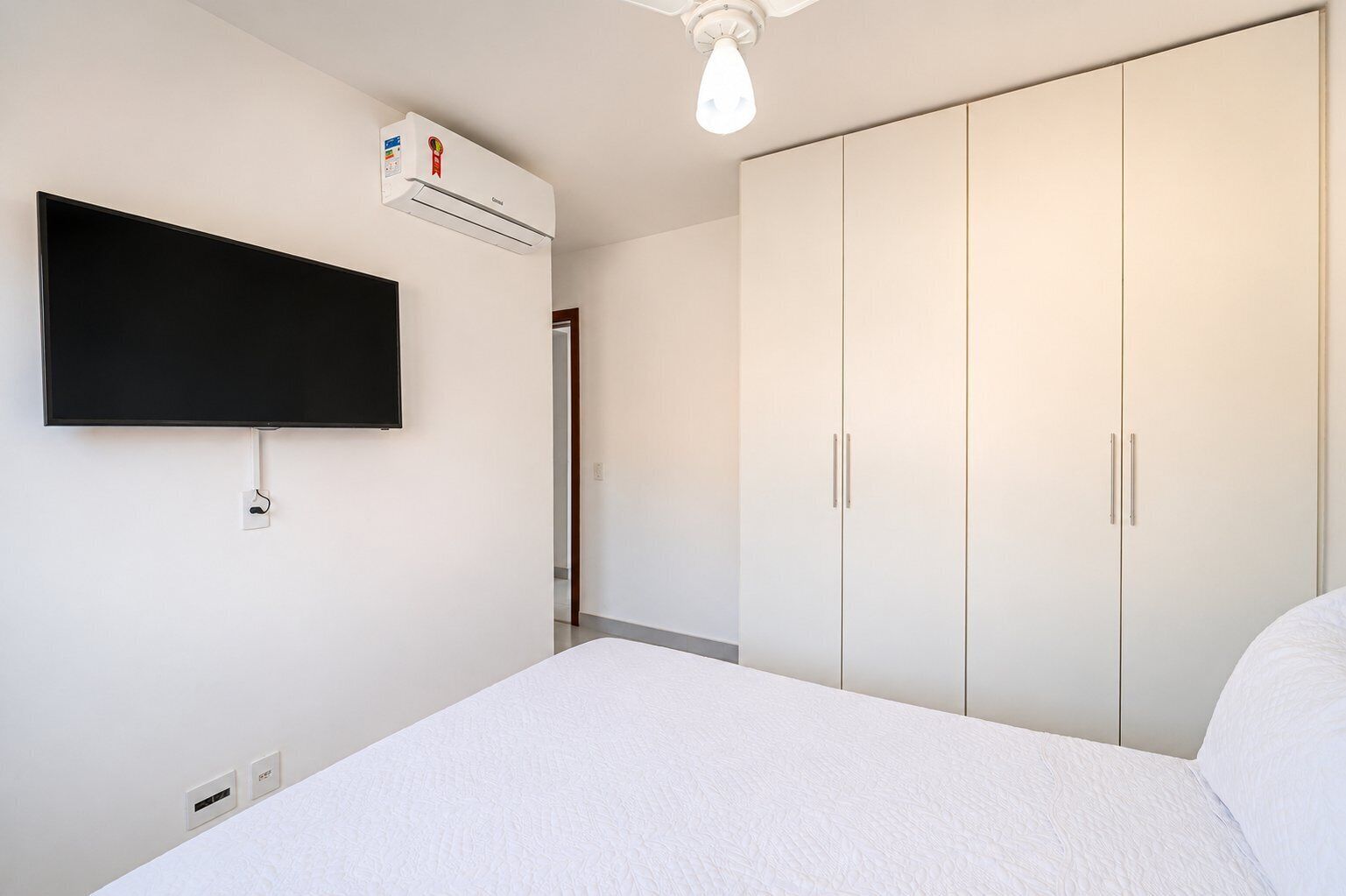 A bedroom with a bed, a flat-screen TV mounted on the wall, a wardrobe, and an air conditioning unit.