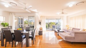 A spacious living area with a sofa, dining table, and chairs, a balcony with a view of greenery, and ceiling fans. - The Wreck Room, 6 Flannel Flower Fairway - fantastic house filled with activities for all (Shoal Bay)