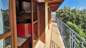 Un balcón de madera con vista a árboles, una zona de cocina con armarios rojos y una puerta de vidrio que da a un espacio interior.