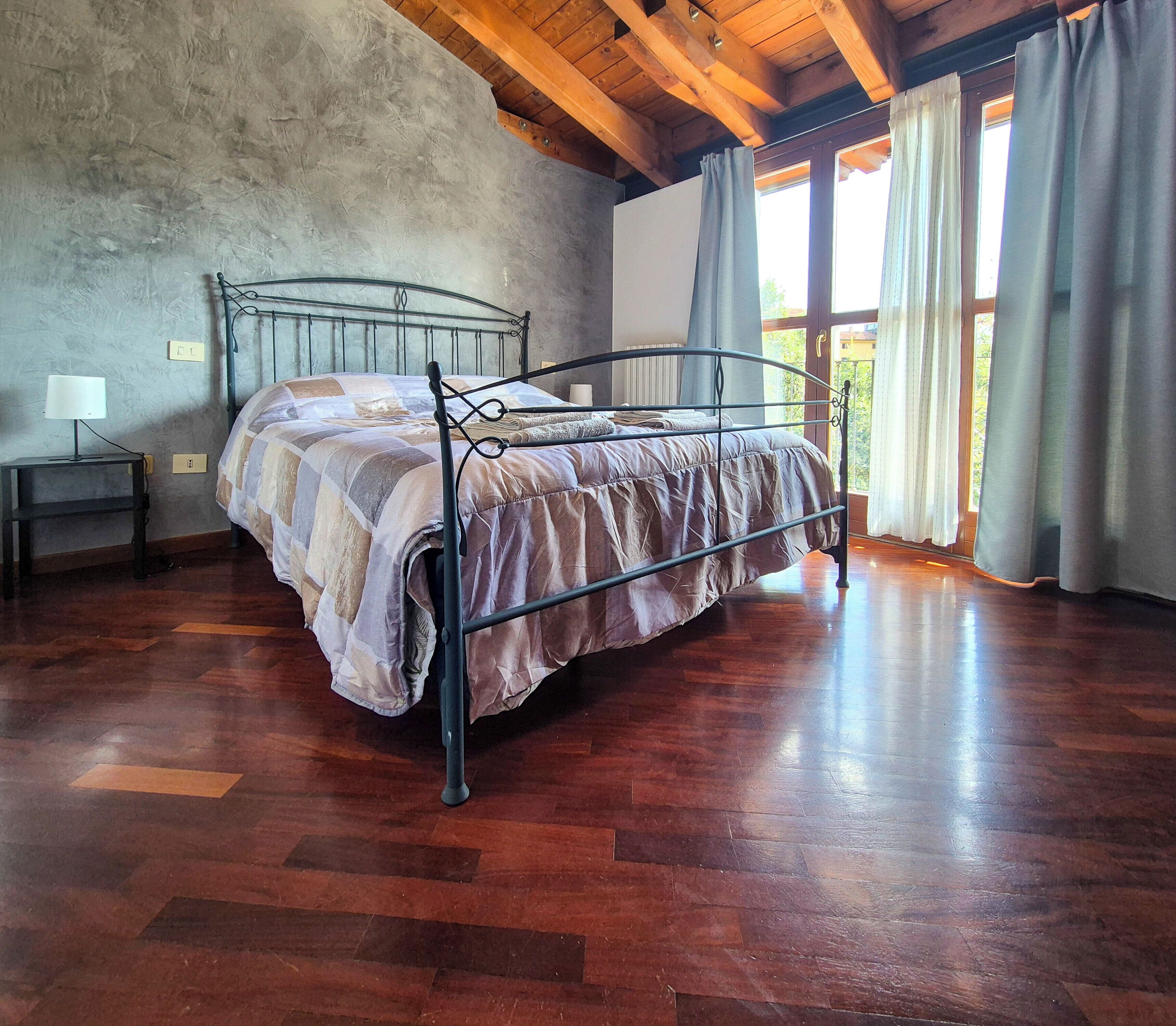 A bedroom with a metal bed frame, wooden flooring, a nightstand with a lamp, and a window with curtains.