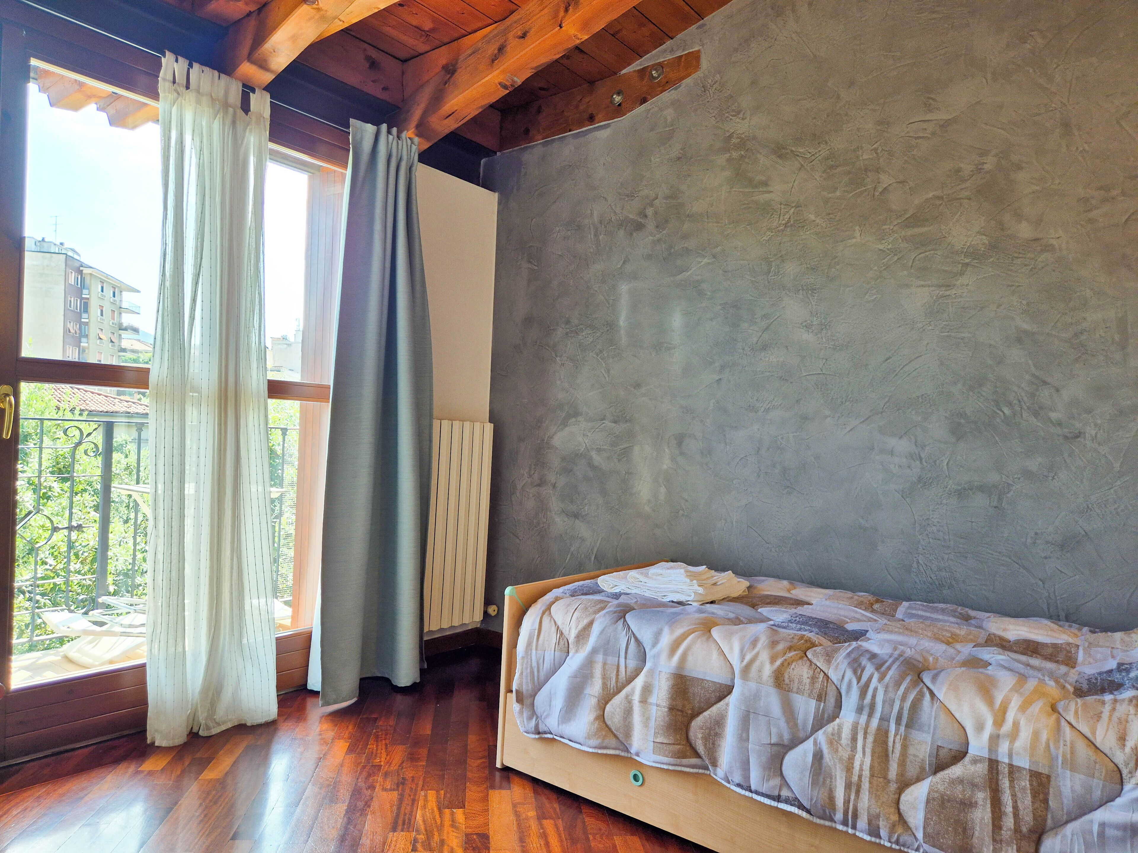 Een slaapkamer met een houten vloer, een bed met een beddegoed met dessin, een raam met gordijnen en uitzicht op groen.